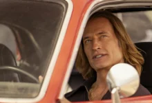 Cena da série inédita "Duster" (2025), da Max, com o ator Josh Holloway dirigindo um carro antigo vermelho; ele interpreta um personagem de visual marcante com cabelos longos loiros e expressão determinada.