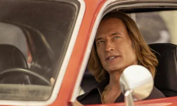 Cena da série inédita "Duster" (2025), da Max, com o ator Josh Holloway dirigindo um carro antigo vermelho; ele interpreta um personagem de visual marcante com cabelos longos loiros e expressão determinada.