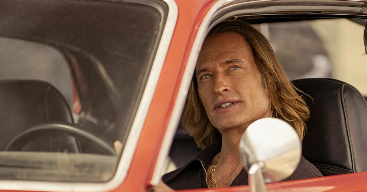 Cena da série inédita "Duster" (2025), da Max, com o ator Josh Holloway dirigindo um carro antigo vermelho; ele interpreta um personagem de visual marcante com cabelos longos loiros e expressão determinada.