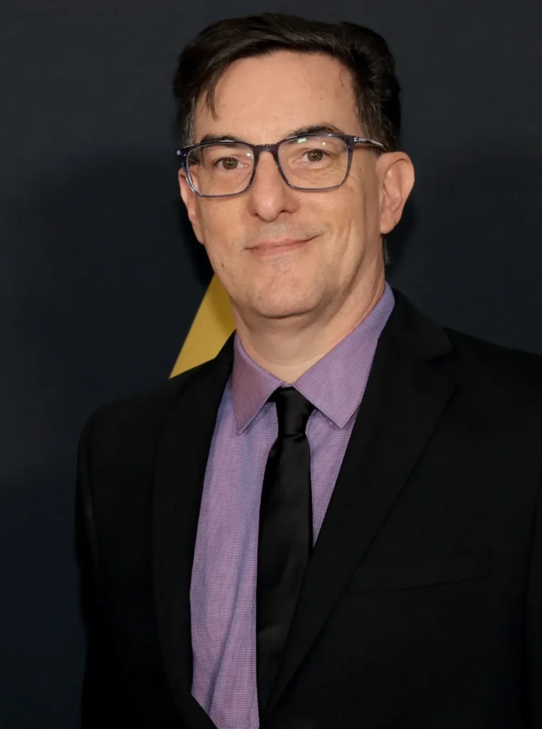 Eric Heisserer, roteirista e criador de séries, em evento de gala usando terno preto, camisa lilás e gravata preta, posando para fotos no tapete vermelho.