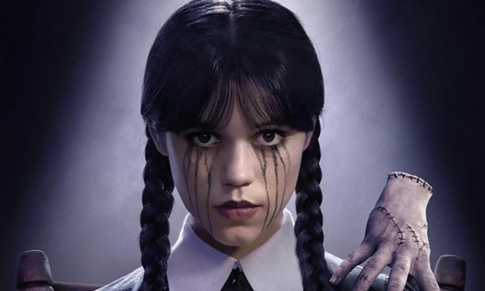Imagem promocional da 2ª temporada de Wandinha, série da Netflix, com a personagem Wandinha Addams (Jenna Ortega) em destaque, com lágrimas negras escorrendo do rosto e a Mãozinha ao seu lado.