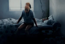 Cena do filme Até a Última Gota, da Netflix, mostra Taraji P. Henson sentada à beira de uma cama em um quarto escuro e desgastado, com expressão de cansaço, enquanto uma criança dorme ao fundo.