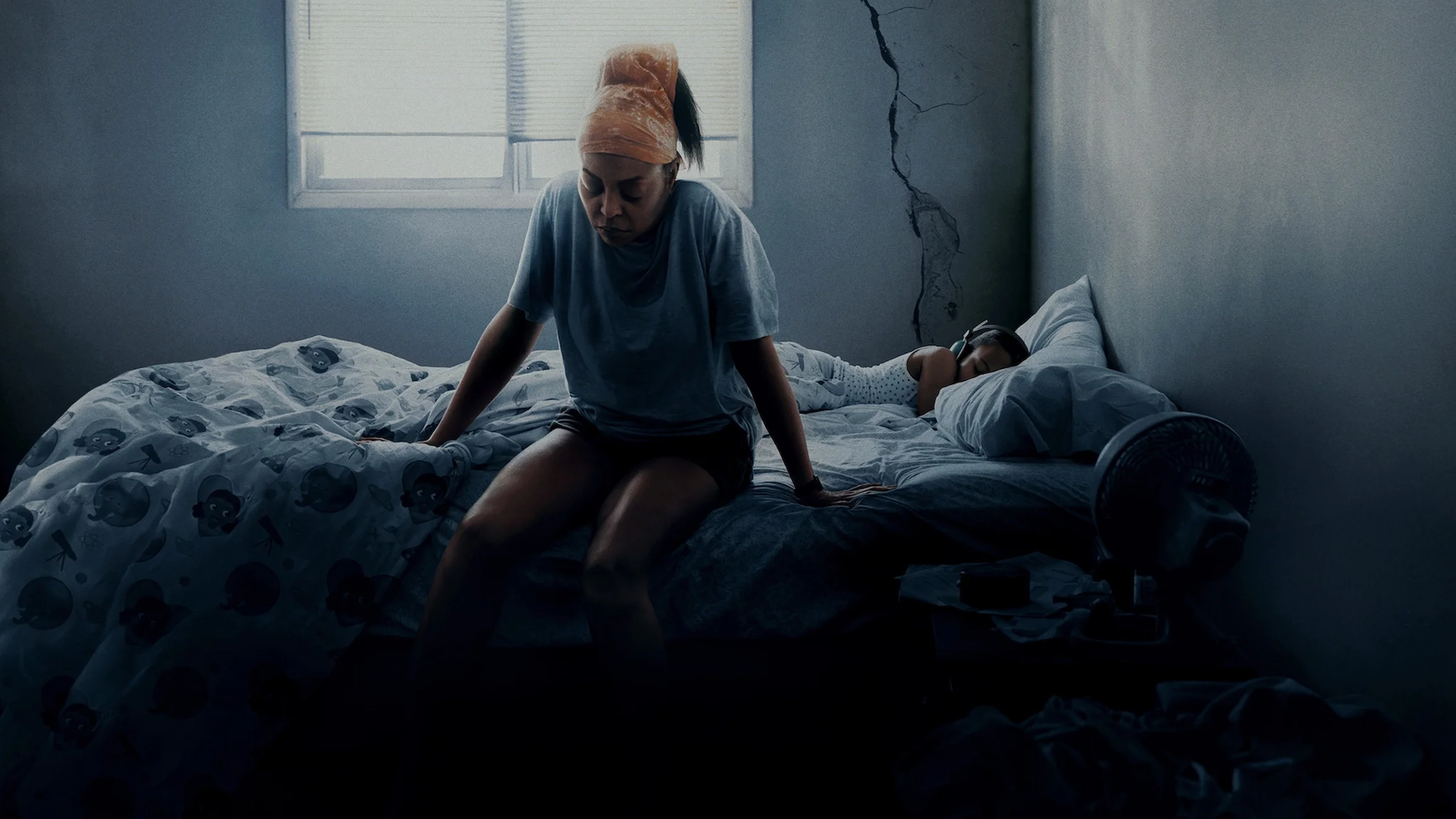 Cena do filme Até a Última Gota, da Netflix, mostra Taraji P. Henson sentada à beira de uma cama em um quarto escuro e desgastado, com expressão de cansaço, enquanto uma criança dorme ao fundo.