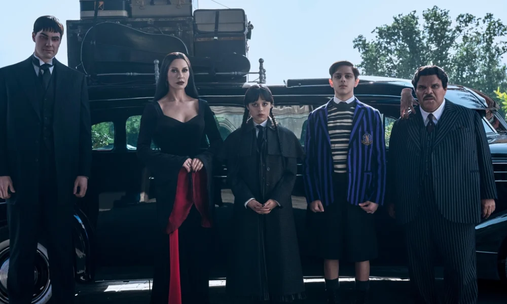 Cena da segunda temporada de Wandinha com Joonas Suotamo, Catherine Zeta-Jones, Jenna Ortega, Isaac Ordonez e Luis Guzmán posando em frente a um carro antigo.