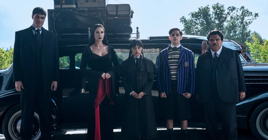 bastidores da 2a temporada de wandinha Cena da segunda temporada de Wandinha com Joonas Suotamo, Catherine Zeta-Jones, Jenna Ortega, Isaac Ordonez e Luis Guzmán posando em frente a um carro antigo.