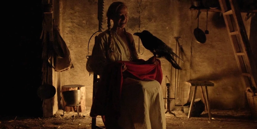 corvo a Kate Dickie como Katherine em A Bruxa (2015), sentada em ambiente sombrio com um corvo pousado em seu colo, em uma das cenas mais inquietantes do terror dirigido por Robert Eggers
