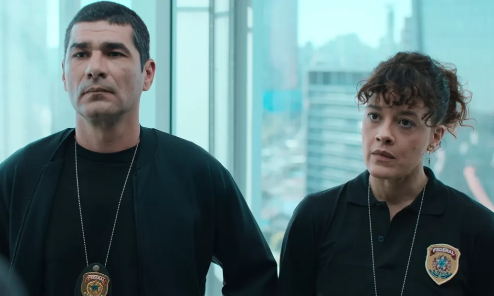 Os atores Rômulo Braga e Maeve Jinkings, como os agentes federais Benício e Suellen, aparecem lado a lado em um escritório envidraçado com vista para a cidade. Ambos vestem roupas escuras com distintivos da Polícia Federal do Brasil, com expressões sérias.