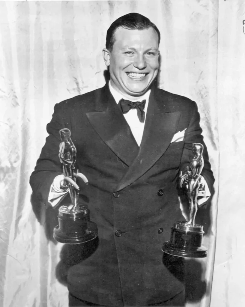harold russell oscars Harold Russell sorrindo enquanto segura dois prêmios Oscar, vestindo smoking, em frente a uma cortina clara. A imagem é em preto e branco.