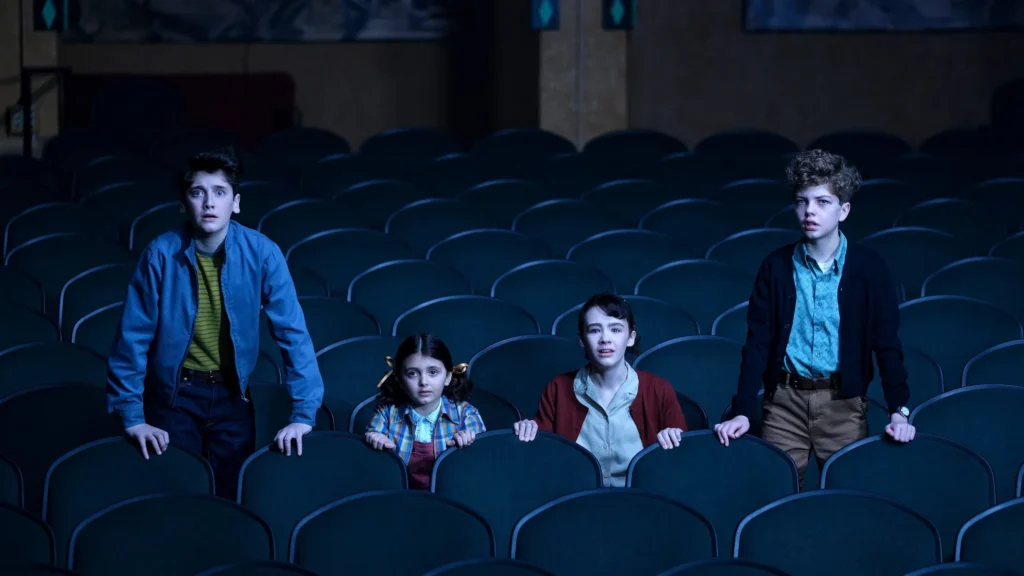 It: Bem-Vindo a Derry (2025) – elenco mirim em momento de tensão Cena da série It: Bem-Vindo a Derry (2025), mostrando os personagens Annie McCormick, Boone Storm, Hunter Storm Baker e Chris Jiggins em um cinema vazio, com expressão de surpresa e tensão, sob iluminação azulada — derivada do universo de terror criado por Stephen King.