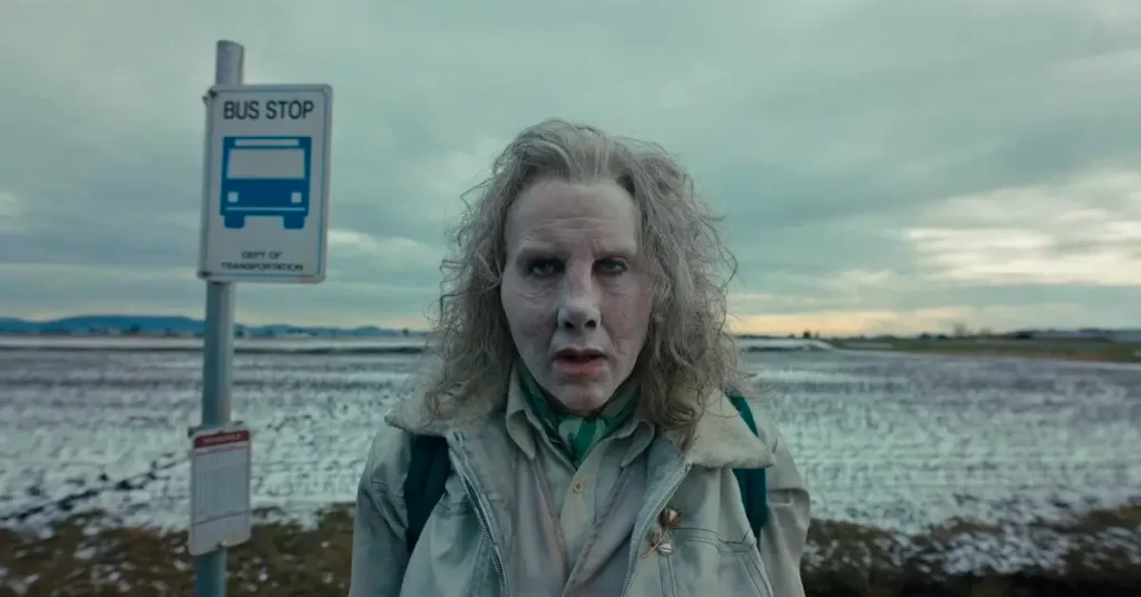 Nicolas Cage maquiado e com expressão perturbadora em cena do filme LongLegs – Vínculo Mortal, diante de uma placa de ponto de ônibus em uma paisagem fria e desolada.