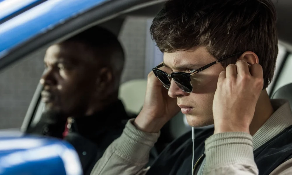 novo trailer de em ritmo de fuga Cena do filme Em Ritmo de Fuga com Ansel Elgort usando óculos escuros e fones de ouvido, enquanto dirige um carro com Jamie Foxx no banco do passageiro.