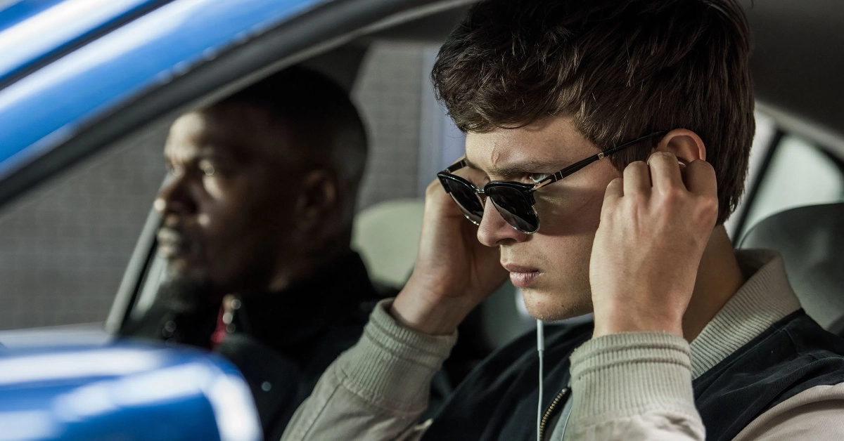 Cena do filme Em Ritmo de Fuga com Ansel Elgort usando óculos escuros e fones de ouvido, enquanto dirige um carro com Jamie Foxx no banco do passageiro.