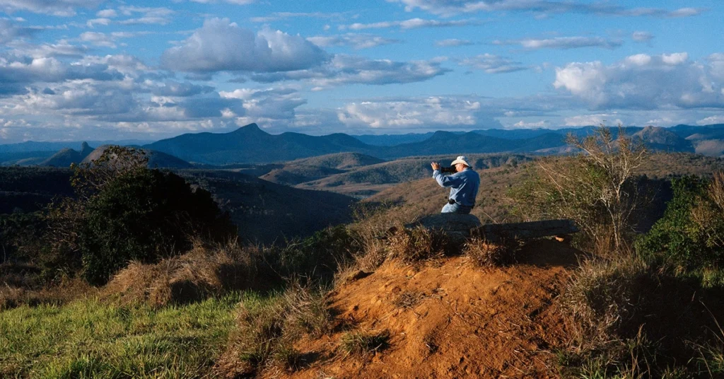 Sebastião Salgado fotografa uma paisagem montanhosa sob céu azul com nuvens, em cena do documentário O Sal da Terra.