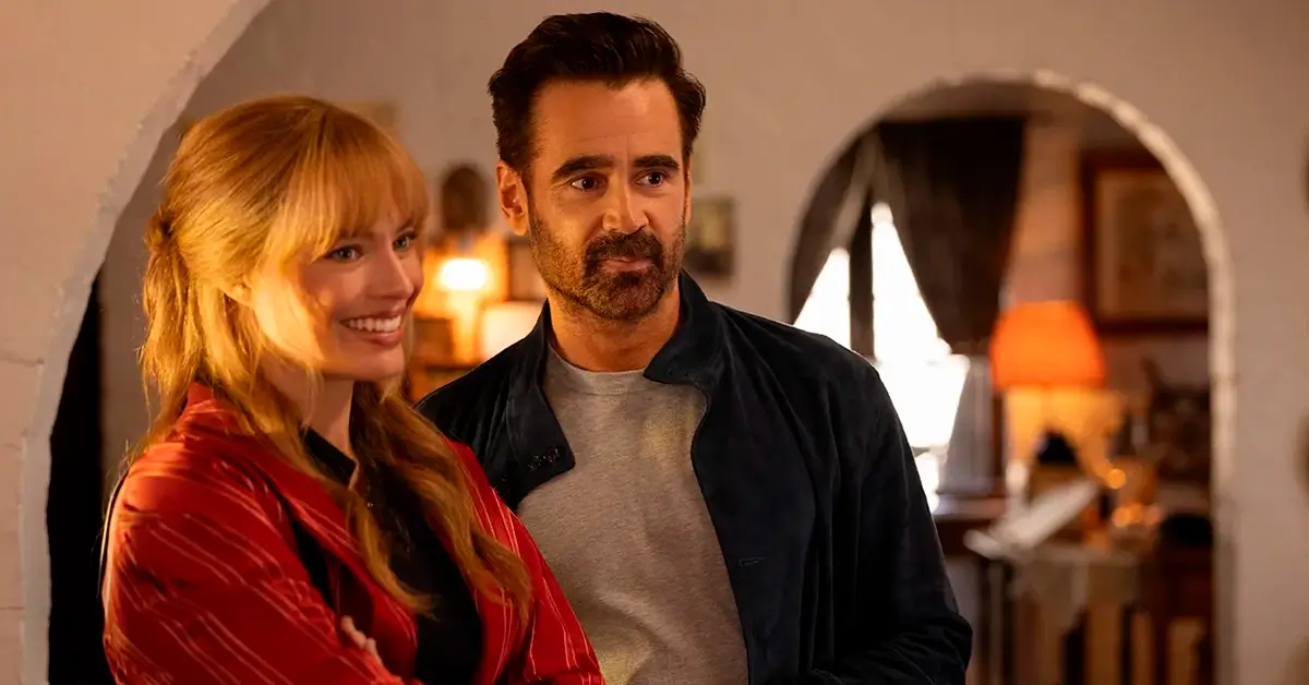 Colin Farrell e Margot Robbie sorrindo em cena do trailer de A Grande Viagem da Sua Vida, com ambiente interno iluminado ao fundo.