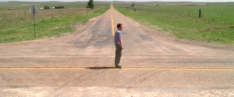 Tom Hanks em cena final de Náufrago (2000), parado em um cruzamento no meio do campo, refletindo sobre seu futuro.