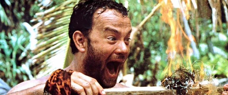 Cena do filme Náufrago (2000) com Tom Hanks comemorando euforicamente após conseguir fazer fogo pela primeira vez na ilha deserta.