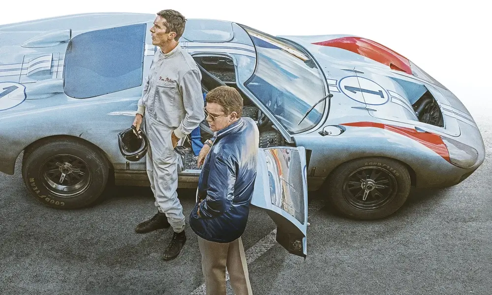 filme sobre f1 ford vs ferrari Christian Bale, vestindo um macacão de corrida claro, em pé ao lado de um Ford GT40 prateado com detalhes em vermelho e azul. Matt Damon, com óculos e uma jaqueta azul escura, está ao lado da porta aberta do carro, observando. O carro de corrida exibe o número "1".