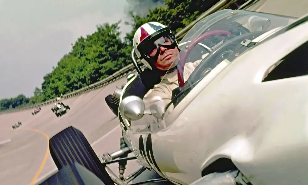 filme sobre f1 grand Close-up de James Garner com capacete tricolor e óculos de proteção, dentro do cockpit de um carro de Fórmula 1, em curva de uma pista com outros carros ao fundo.