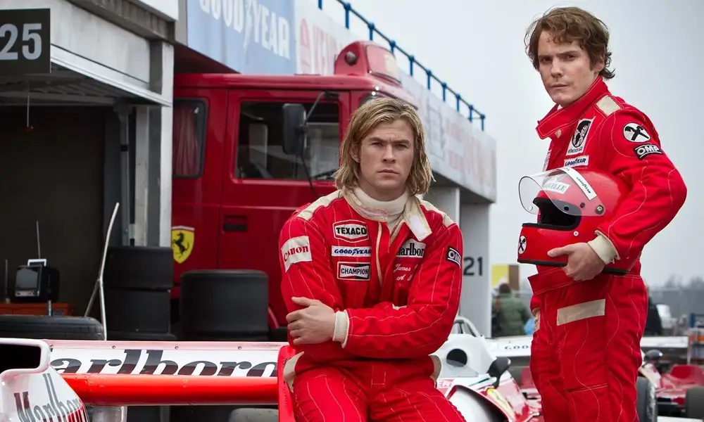 filme sobre f1 rush no limite da emocao Chris Hemsworth e Daniel Brühl caracterizados como James Hunt e Niki Lauda em um pit lane. Hemsworth está sentado ao lado de um carro de Fórmula 1 vermelho e branco; Brühl está em pé com um capacete, ambos com macacões da Ferrari.