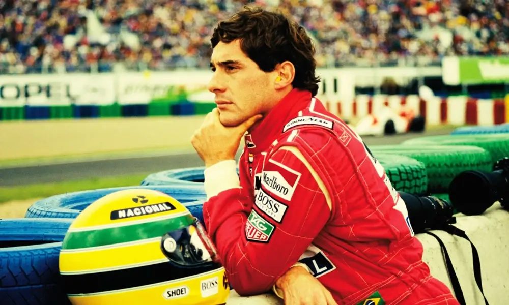 filme sobre f1 senna 2010 Ayrton Senna em close-up de perfil, vestindo macacão vermelho com expressão pensativa e seu capacete ao lado. Cena do documentário Senna (2010), sobre a vida do piloto.