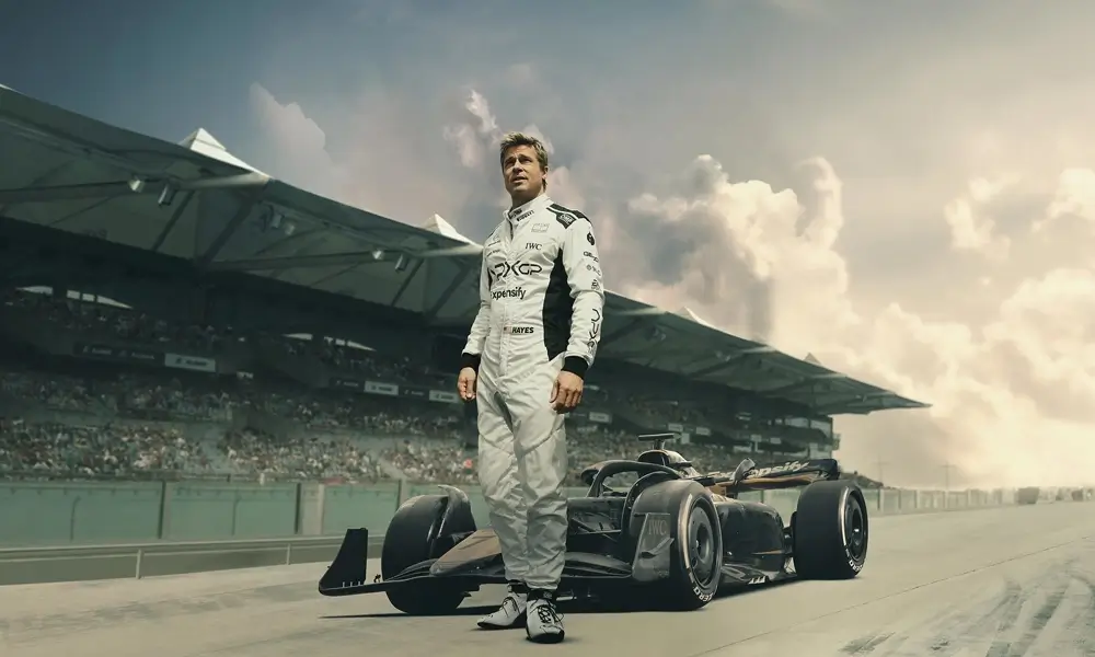 filme sobre formula 1 Brad Pitt como o piloto Sonny Hayes ao lado de um carro de Fórmula 1 moderno em uma pista de corrida, com arquibancadas e céu nublado ao fundo.