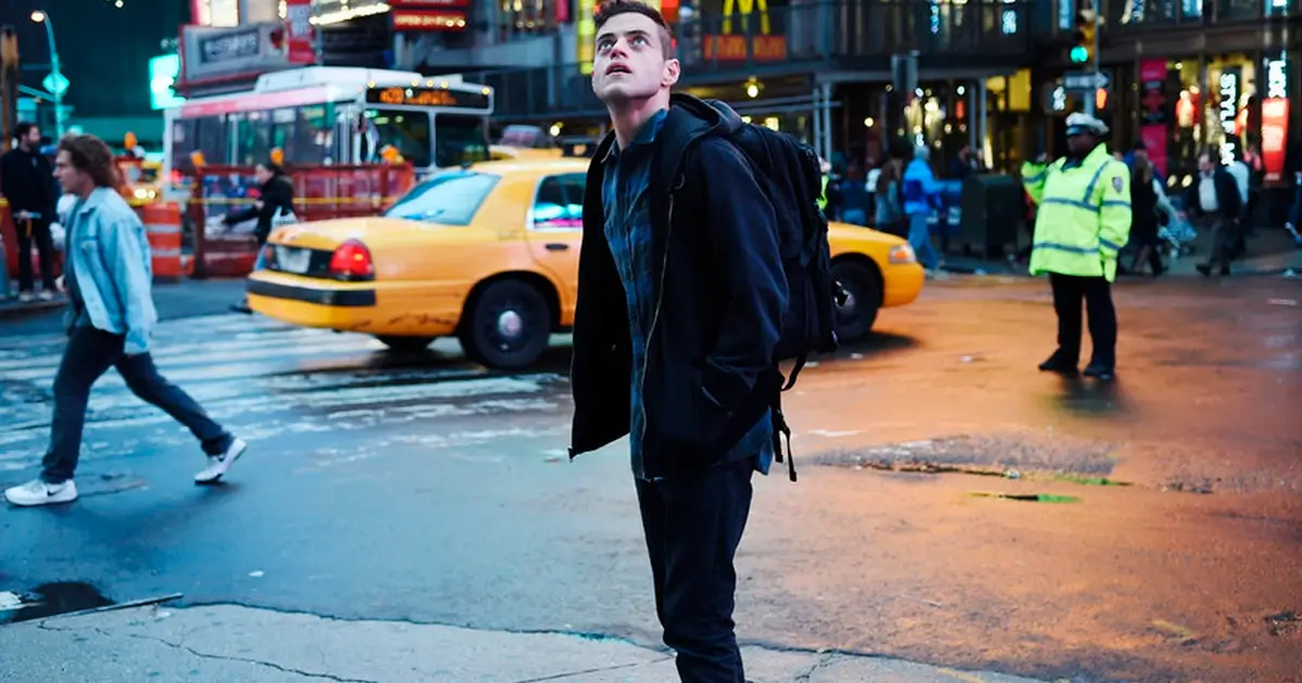 Elliot em cena noturna do episódio piloto de Mr. Robot Elliot (Rami Malek) em cena noturna do primeiro episódio de Mr. Robot, observando a cidade iluminada enquanto táxis e pedestres passam.