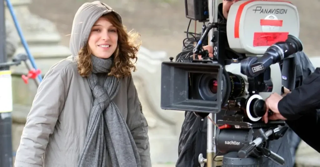 natalie portman diretora Natalie Portman no set de filmagem, vestida com casaco e cachecol, ao lado de uma câmera cinematográfica.