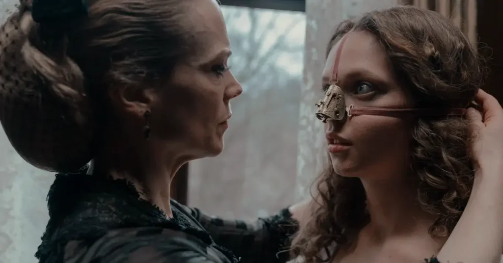 Cena do filme The Ugly Stepsister (2025) com Lea Myren usando máscara metálica no rosto e Ane Dahl Torp ajustando o acessório