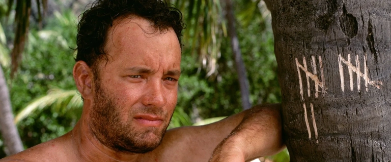Tom Hanks como Chuck Noland sentado ao lado de uma árvore marcada com riscos para contar os dias, em cena do filme Náufrago (2000).