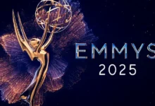 Uma imagem da estatueta dourada do Emmy, com braços estendidos segurando um globo, cercada por brilhos dourados e azuis que se assemelham a fogos de artifício ou explosões de luz. À direita, as palavras "EMMYS 2025" estão escritas em letras maiúsculas, com "EMMYS" em azul metálico e "2025" em branco. O fundo é um céu noturno escuro.