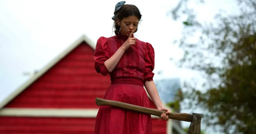 Mia Goth como Pearl segurando um machado em frente a uma casa vermelha, filme de terror Pearl.