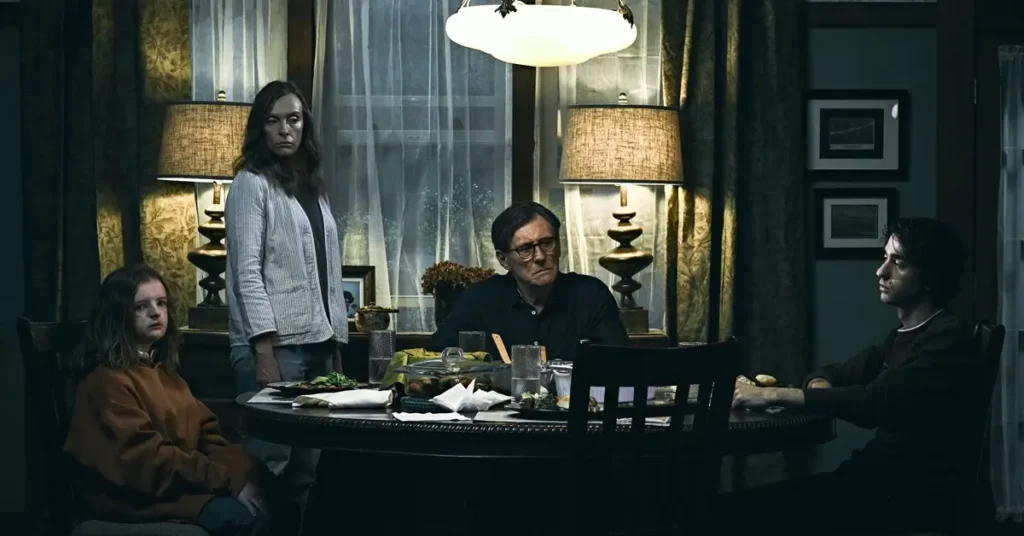 hereditario elenco principal Toni Collette, Gabriel Byrne, Alex Wolff e Milly Shapiro em uma cena dramática do filme Hereditário (2018), reunidos em volta da mesa de jantar