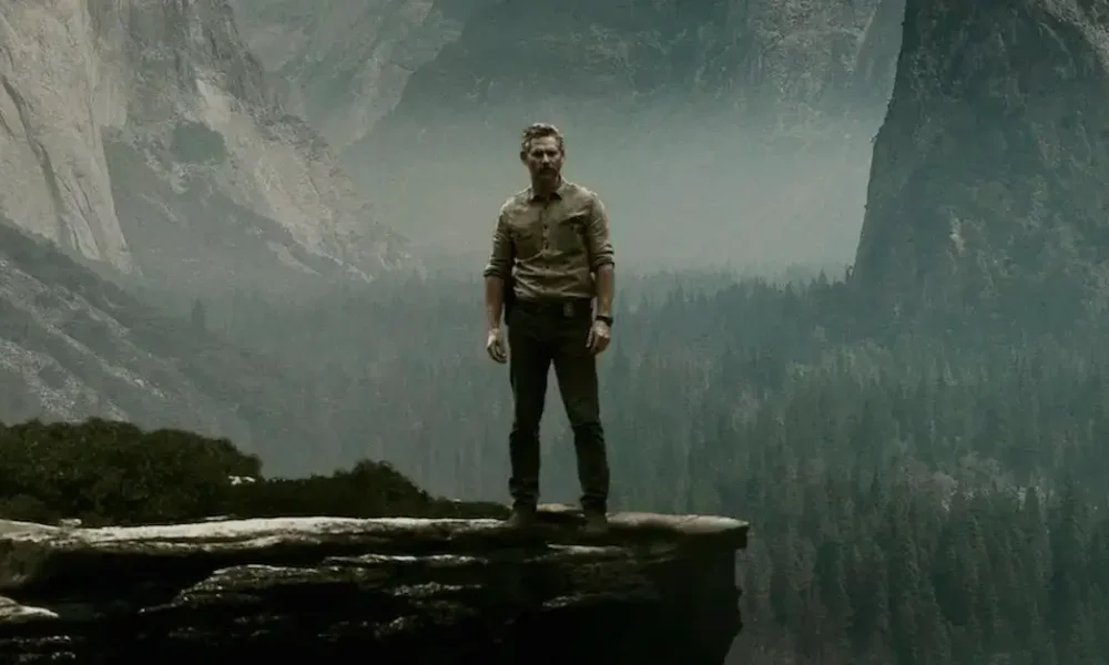 Eric Bana em pé na beira de um penhasco com uma floresta e montanhas ao fundo na minissérie Indomável (2025), da Netflix.