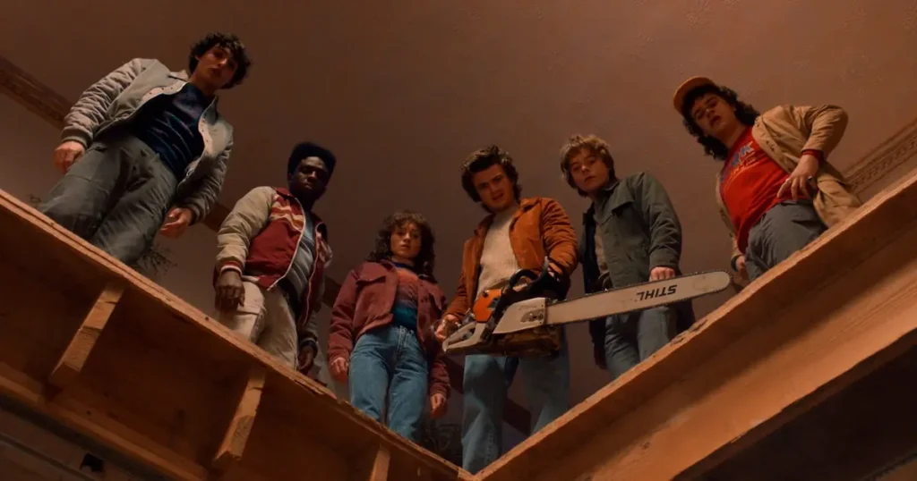  Os personagens de "Stranger Things" Lucas Sinclair, Nancy Wheeler, Steve Harrington, Jonathan Byers e Dustin Henderson olham para baixo com expressões tensas, enquanto Steve segura uma motosserra Stihl, prenunciando desafios perigosos na quinta e última temporada da série da Netflix.