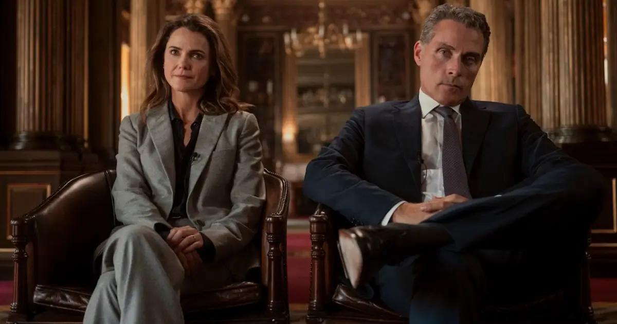 Keri Russell e Rufus Sewell sentados em poltronas formais em cena da terceira temporada de A Diplomata.