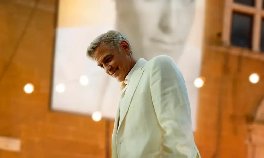 George Clooney em cena do filme Jay Kelly (2025), vestindo terno claro em ambiente interno com iluminação cinematográfica