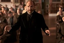Cena do filme "Sinfonia de Guerra (The Choral)" com Ralph Fiennes em primeiro plano, cercado por uma plateia atenta.