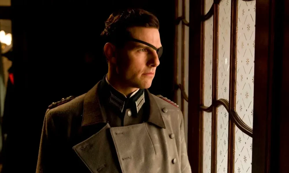 Tom Cruise como Coronel Claus von Stauffenberg em Operação Valquíria - homem de perfil usando uniforme militar cinza com insígnias, óculos e tapa-olho, olhando pensativamente pela janela em ambiente interno com iluminação quente