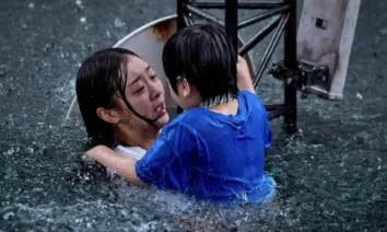 Mulher assustada segura uma criança enquanto tenta se manter à tona em meio a uma enchente, com destroços metálicos ao fundo, cena do filme sul-coreano A Grande Inundação (The Great Flood) da Netflix.