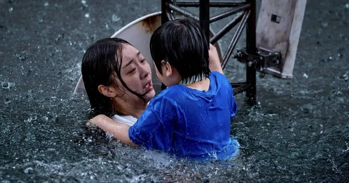 Mulher assustada segura uma criança enquanto tenta se manter à tona em meio a uma enchente, com destroços metálicos ao fundo, cena do filme sul-coreano A Grande Inundação (The Great Flood) da Netflix.