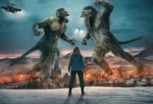 Cena épica de O Troll da Montanha 2, com dois trolls gigantes lutando diante de uma cidade em chamas e Ine Marie Wilmann observando de costas.