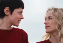 Emma Corrin e Maika Monroe se encaram em cena do filme A Centésima Noite (2025), vestindo roupas vermelhas sob um céu nublado.