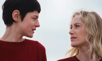 Emma Corrin e Maika Monroe se encaram em cena do filme A Centésima Noite (2025), vestindo roupas vermelhas sob um céu nublado.