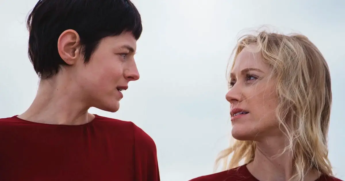 Emma Corrin e Maika Monroe se encaram em cena do filme A Centésima Noite (2025), vestindo roupas vermelhas sob um céu nublado.