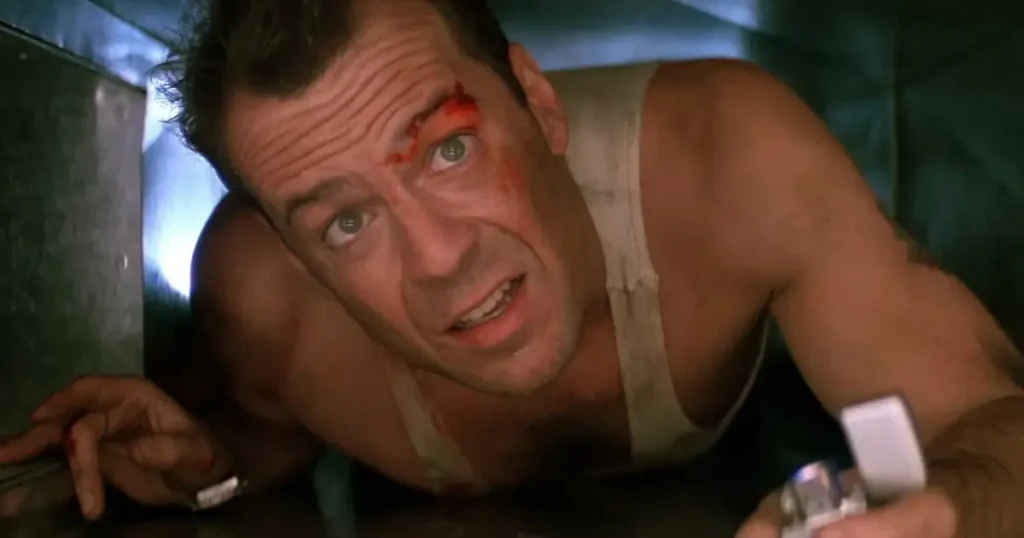 duro de matar Bruce Willis como John McClane rastejando pelo duto de ventilação em Duro de Matar, ferido e tenso na sequência de ação ambientada na véspera de Natal.