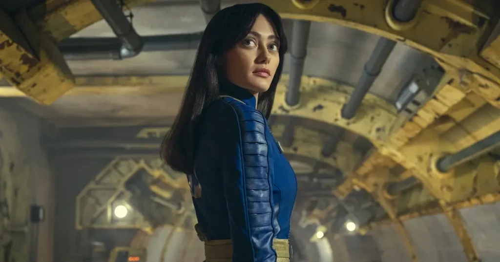 Lucy MacLean em traje do Vault 33 dentro de um corredor subterrâneo no episódio “Os Necróticos”, da série Fallout (2024).
