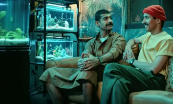 Atores Mohammed Aldokhei e Abdullah AlDaris em cena do filme Sequestro Fake (2025), sentados em uma sala iluminada por aquários.