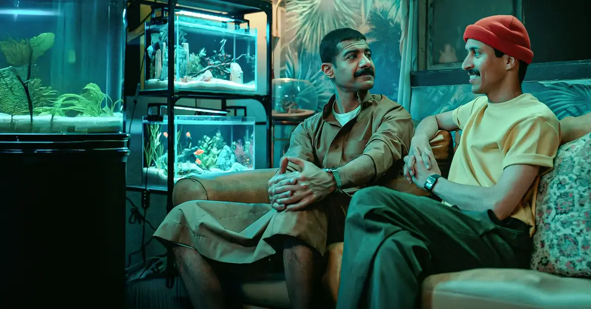 Atores Mohammed Aldokhei e Abdullah AlDaris em cena do filme Sequestro Fake (2025), sentados em uma sala iluminada por aquários.