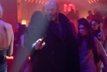 Jason Statham armado em cena do filme Shelter (2026), dirigido por Ric Roman Waugh, dentro de uma boate iluminada.