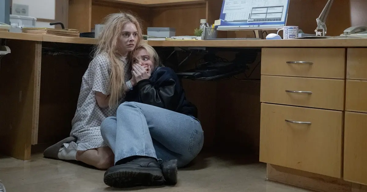 Kathryn Newton e Samara Weaving escondidas sob uma mesa em cena tensa de Casamento Sangrento: A Viúva (2026), do Trailer de Casamento Sangrento: A Viúva.